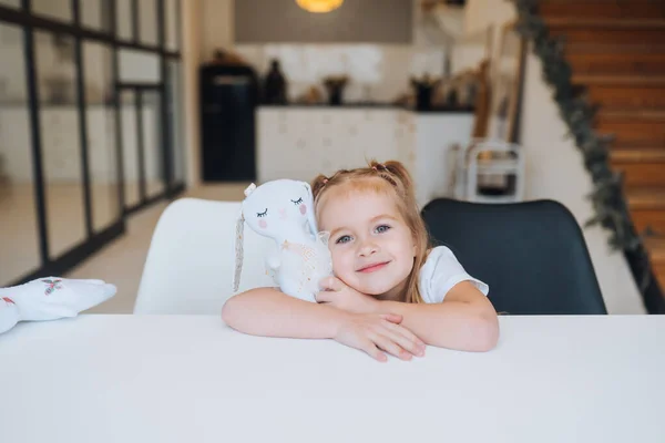 Kleines lächelndes Mädchen umarmt Lieblingsspielzeug, während es am Tisch sitzt — Stockfoto