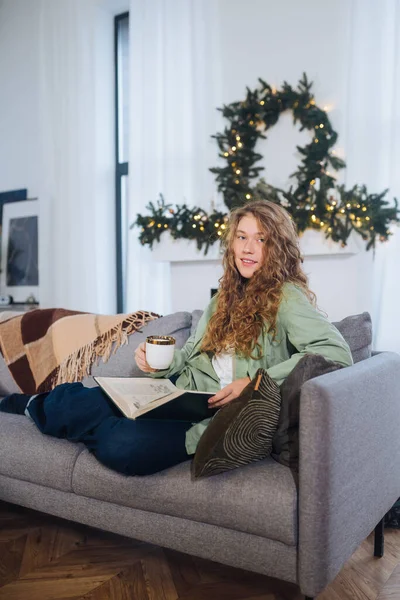 Wanita muda di sofa membaca buku dan menikmati kopi — Stok Foto