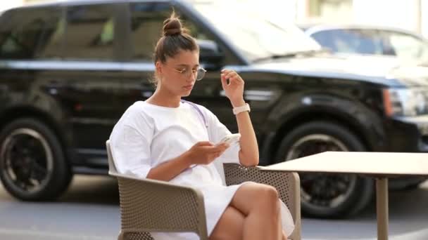 Piękna młoda kobieta siedzi w ulicznej kawiarni za pomocą smartfona — Wideo stockowe