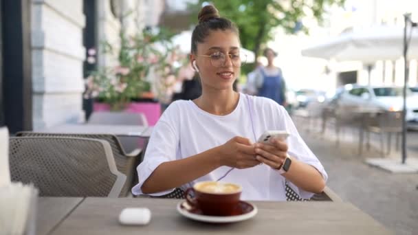 Hermosa joven sentada en la cafetería de la calle usando un teléfono inteligente — Vídeo de stock