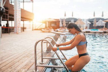 Mayo giymiş genç bir kadın havuzdan çıkmak için merdivene tırmanıyor.