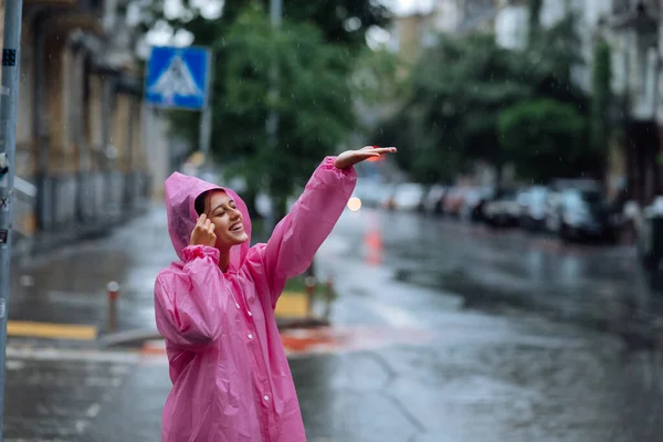 Young smiling woman with a pink raincoat on the street while enjoying a walk through the city on a rainy day.