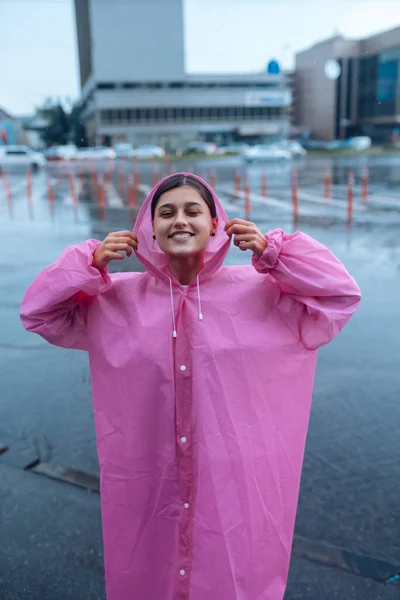 Young smiling woman with a pink raincoat on the street while enjoying a walk through the city on a rainy day.