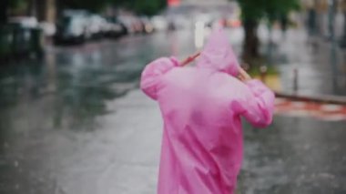 Yağmurlu bir günde şehirde yürürken sokakta pembe yağmurluklu gülümseyen genç bir kadın..
