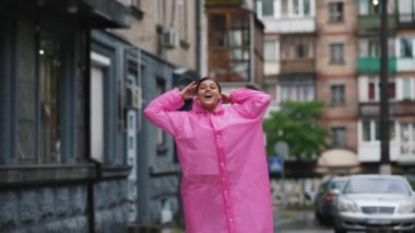 Yağmurlu bir günde şehirde yürürken sokakta pembe yağmurluklu gülümseyen genç bir kadın..