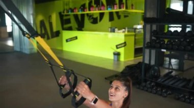 Spor kıyafetleri içinde mükemmel vücutlu genç atletik bir kadın spor salonunda trx fitness kayışlarıyla egzersiz yapıyor.