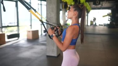 Spor kıyafetleri içinde mükemmel vücutlu genç atletik bir kadın spor salonunda trx fitness kayışlarıyla egzersiz yapıyor.