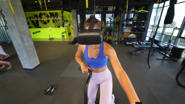 在一个宽敞的健身房里 坐在跑车上 戴着一副虚拟现实的眼镜 微笑着美丽的黑发 — 图库视频影像