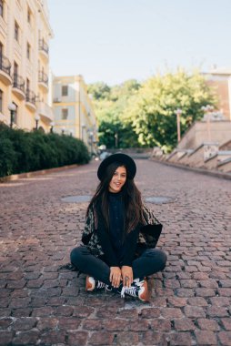 Şapkalı genç bir kadın şehir parke taşında oturuyor, ılık bir sonbahar günü.