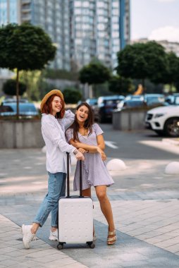 Elinde bavul olan iki genç kadın sokakta kameraya poz veriyor.