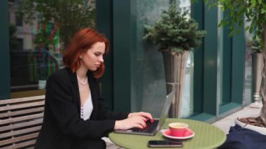 Güzel kızıl saçlı kadın dışarıda, kafede dizüstü bilgisayarda çalışıyor.