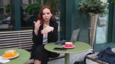 Neşeli, zarif giyimli kadın kameraya bakarken ve konuşurken gülümsüyor kaldırımdaki kafede dizüstü bilgisayarla oturuyor.