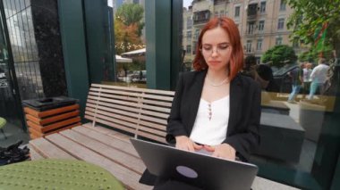 Güzel kızıl saçlı kadın dışarıda, kafede dizüstü bilgisayarda çalışıyor.