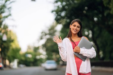 Dizüstü bilgisayar tutan genç bir kadın sokaktaki kameraya bakıyor.