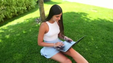 Uzaktan kumanda konsepti. Genç bir kız parktaki temiz havada dizüstü bilgisayarla çimlerin üzerinde oturuyor..