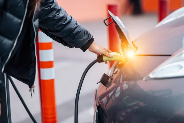 充電ステーションに停車中の電気自動車で移動する若い美しい女性充電中に喜んで笑顔スマートフォンでインターネットを接続ケーブルを接続スタンド — ストック写真