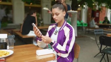 Genç bir kadın terasta, kafede oturmuş elinde akıllı telefonuyla çay ya da kahve içiyor.