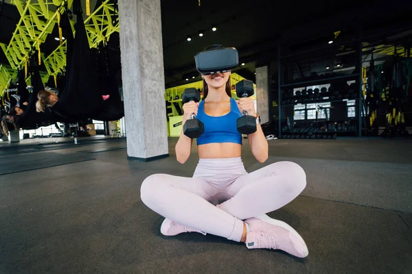 Portrait view of the woman wearing glasses of the virtual reality exercising with dumbbells at gym.