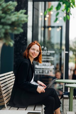 Güzel kızıl saçlı kadın dışarıda, kafede dizüstü bilgisayarda çalışıyor.