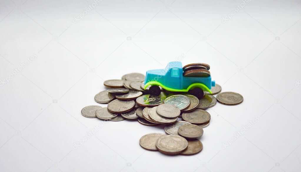 A blue-green truck carrying coins climbs on a pile of coins on a white background.