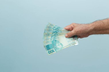  Mavi arka planda 100 Reais banknotla yaşlı bir adamın eli. Ödeme konsepti. Açık mavi arkaplan.