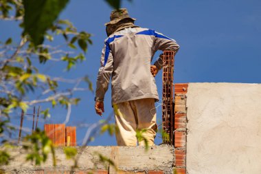 Etrafında ağaç dalları ve arka planında mavi gökyüzü olan bir binanın tepesinde bir tuğla ustası..