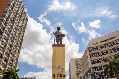 Goiania 'daki Bandeirante Meydanı' nın ortasında, etrafında binalar ve arka planda bulutlu bir gökyüzü olan heykelin fotoğrafı. Praa do Bandeirante.