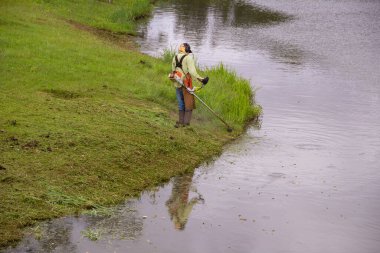 Uygun ekipmanlarla gölün kenarında çim biçen bir işçi..
