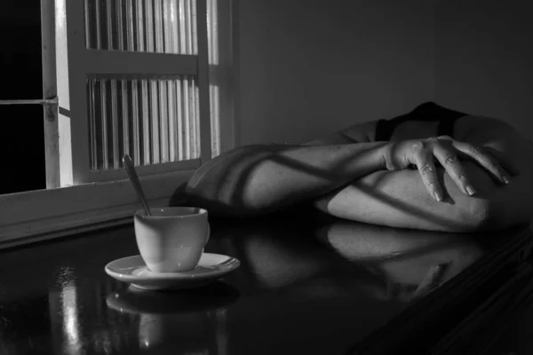 A person with his head down and leaning on a table with a cup of coffee, next to a window. Sadness and tiredness concept. Black and white image.