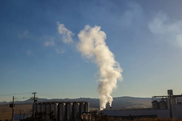 A factory emitting smoke into the environment, on a clear morning with the blue sky.