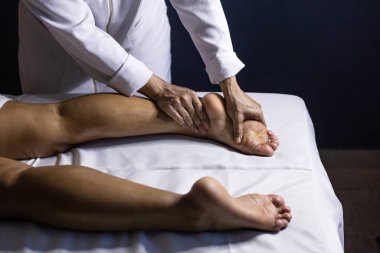 Detail of masseuse hands applying therapeutic massage on the foot of a patient who is lying on a stretcher with white sheet.