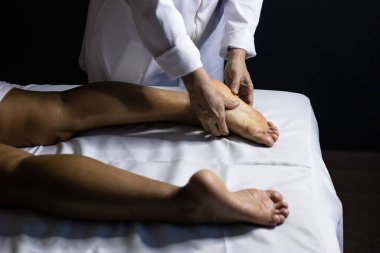 Detail of masseuse hands applying therapeutic massage on the foot of a patient who is lying on a stretcher with white sheet.