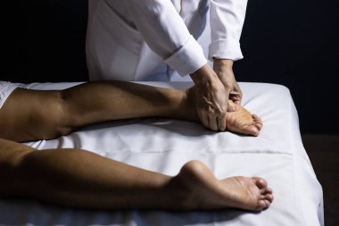 Detail of masseuse hands applying therapeutic massage on the foot of a patient who is lying on a stretcher with white sheet.