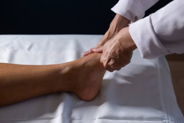 Detail of masseuse hands applying therapeutic massage on the foot of a patient who is lying on a stretcher with white sheet.
