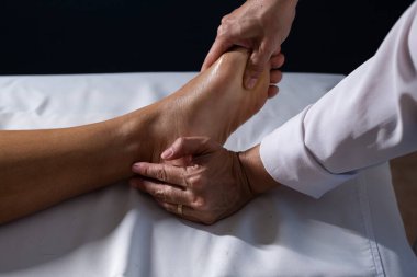 Detail of masseuse hands applying therapeutic massage on the foot of a patient who is lying on a stretcher with white sheet.