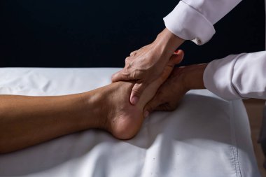 Detail of masseuse hands applying therapeutic massage on the foot of a patient who is lying on a stretcher with white sheet.