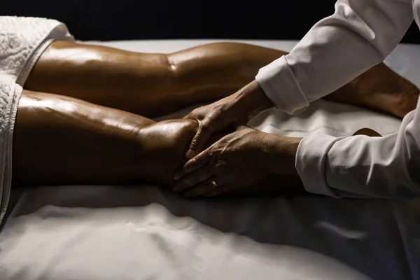 A therapist dressed in white, massaging the leg of a patient lying on a stretcher, using therapeutic oil.