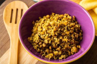 A crockery bowl with brown rice and lentils on a wooden surface, with a wooden spoon and a yellow cloth in the background.