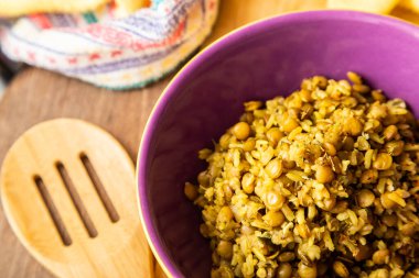 A crockery bowl with brown rice and lentils on a wooden surface, with a wooden spoon and a yellow cloth in the background.