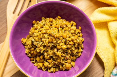 A crockery bowl with brown rice and lentils on a wooden surface, with a wooden spoon and a yellow cloth in the background.