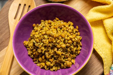 A crockery bowl with brown rice and lentils on a wooden surface, with a wooden spoon and a yellow cloth in the background.