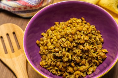 A crockery bowl with brown rice and lentils on a wooden surface, with a wooden spoon and a yellow cloth in the background.