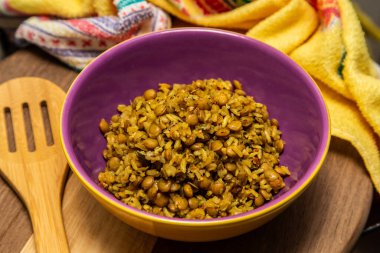 A crockery bowl with brown rice and lentils on a wooden surface, with a wooden spoon and a yellow cloth in the background.