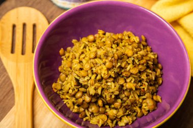 A crockery bowl with brown rice and lentils on a wooden surface, with a wooden spoon and a yellow cloth in the background.