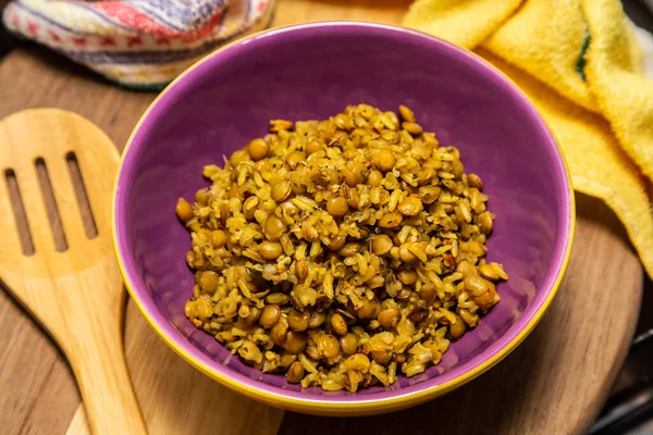 A crockery bowl with brown rice and lentils on a wooden surface, with a wooden spoon and a yellow cloth in the background.