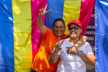 A lesbian couple smiling, hugging, with rainbow flags in the background. Photo taken from the 2022 LGBTQIA+ Pride Parade in the city of Goiania.