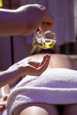 Masaj terapisti, arka planda sedyede yatan bir hastayla ellere yağ döküyor..