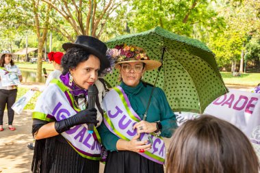Kadın hakları savunucusu gibi giyinmiş, mikrofona konuşan ve oy vermeye teşvik etmek için şemsiye tutan bir kadın..