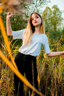 Açık gri tişört ve siyah kot pantolon giyen genç ve güzel bir kız sıcak sonbaharın başlarında yüksek yeşil ve sarı çimlere poz veriyor. Güzel bir kadın modelin açık hava portresi. Dikey çekim.