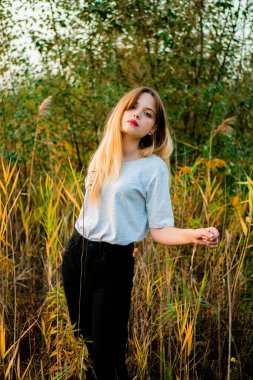 Açık gri tişört ve siyah kot pantolon giyen genç ve güzel bir kız sıcak sonbaharın başlarında yüksek yeşil ve sarı çimlere poz veriyor. Güzel bir kadın modelin açık hava portresi. Dikey çekim.